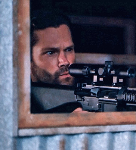 A Man With A Beard Is Holding A Sniper Rifle Behind A Window GIF