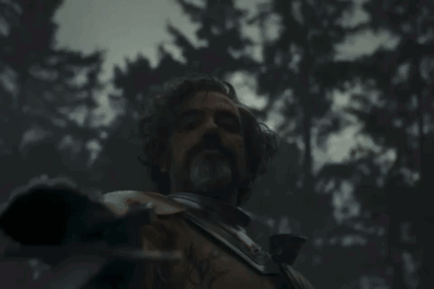 A Man With A Beard Is Holding A Sword In A Dark Forest GIF