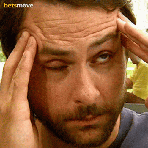 A Man With A Beard Is Holding His Head While Looking At The Camera . GIF