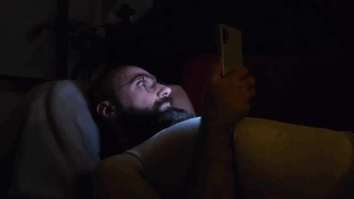 A Man With A Beard Is Laying In Bed Using A Cell Phone GIF