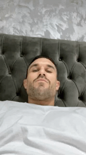 A Man With A Beard Is Laying In Bed With His Eyes Closed And His Head On The Headboard . GIF