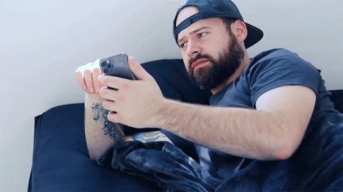 A Man With A Beard Is Laying On A Bed Looking At A Cell Phone GIF