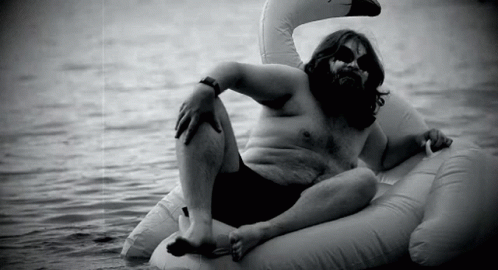 A Man With A Beard Is Laying On An Inflatable Swan In The Water . GIF