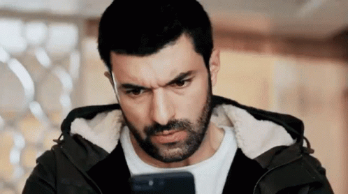 A Man With A Beard Is Looking At His Cell Phone And Making A Funny Face . GIF