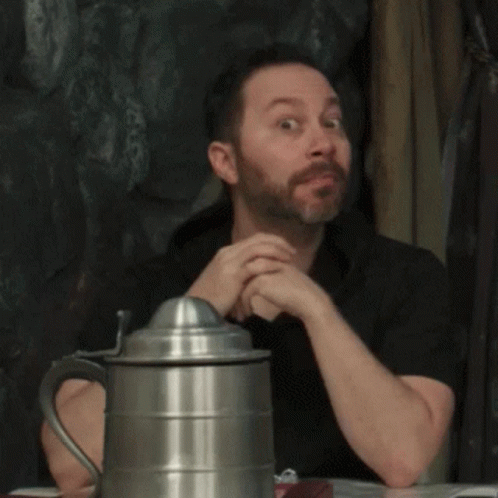 A Man With A Beard Is Sitting At A Table Next To A Metal Pitcher GIF
