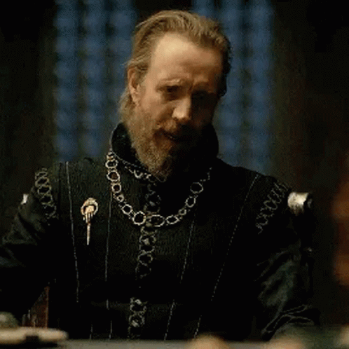 a man with a beard is sitting at a table wearing a black jacket and a gold chain around his neck .