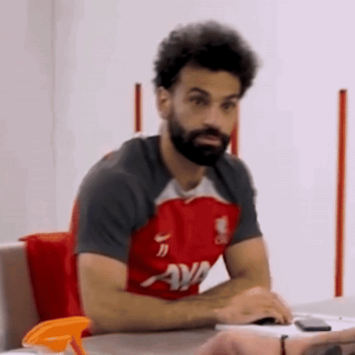 A Man With A Beard Is Sitting At A Table Wearing A Red Shirt That Says Ava On It GIF