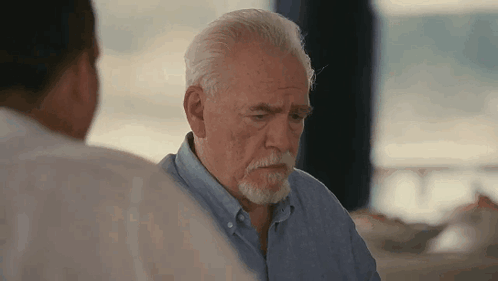 A Man With A Beard Is Sitting At A Table With Another Man . GIF