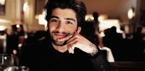 A Man With A Beard Is Sitting At A Table With His Hand On His Chin And Smiling At The Camera . GIF