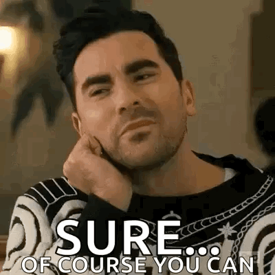 A Man With A Beard Is Sitting At A Table With His Hand On His Chin And The Words `` Sure Of Course You Can '' . GIF