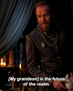 a man with a beard is sitting at a table with the words " my grandson is the future of the realm " above him