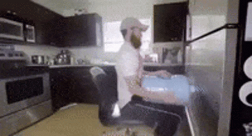 A Man With A Beard Is Sitting In A Chair In A Kitchen Holding A Bucket . GIF