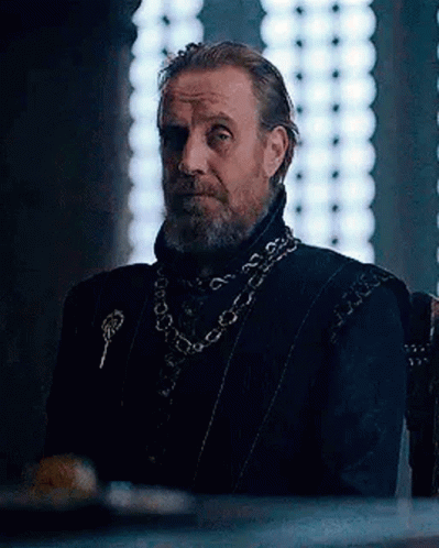 a man with a beard is sitting in a chair in front of a window wearing a chain around his neck .