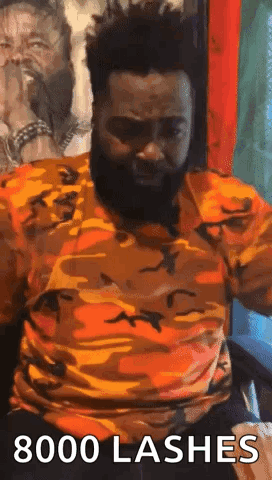A Man With A Beard Is Sitting In A Chair With His Arms Outstretched And A Camouflage Shirt On . GIF