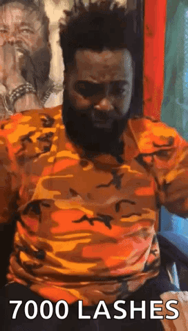 A Man With A Beard Is Sitting In A Chair With His Arms Outstretched And A Camouflage Shirt On . GIF