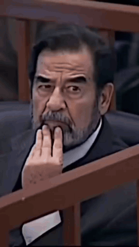 A Man With A Beard Is Sitting In A Chair With His Hand On His Chin And Looking At The Camera . GIF