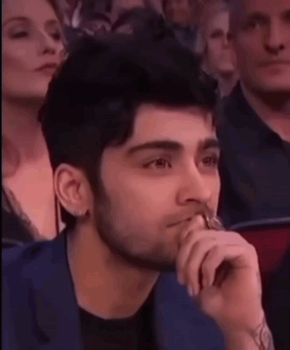 A Man With A Beard Is Sitting In A Crowd With His Hand To His Mouth . GIF