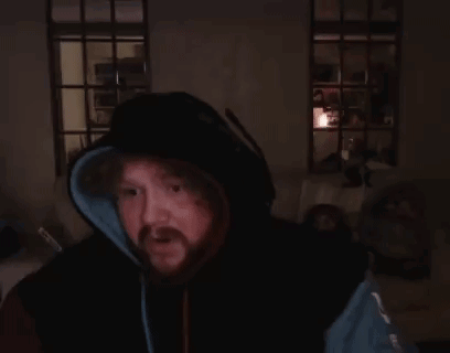 A Man With A Beard Is Sitting In A Dark Room With His Hands Folded . GIF