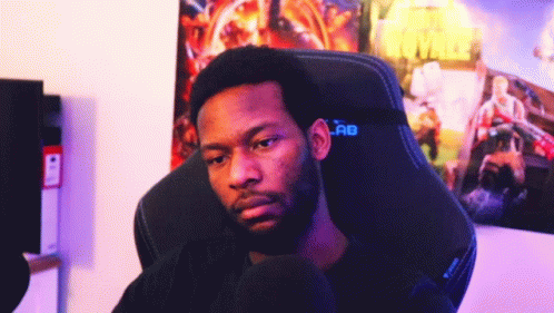 A Man With A Beard Is Sitting In A Gaming Chair In Front Of A Poster Of Battle Royale GIF