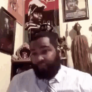 A Man With A Beard Is Sitting In A Room With A Lot Of Paintings On The Wall . GIF