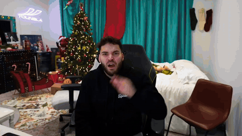 A Man With A Beard Is Sitting In Front Of A Christmas Tree And A Sign That Says Younbla GIF