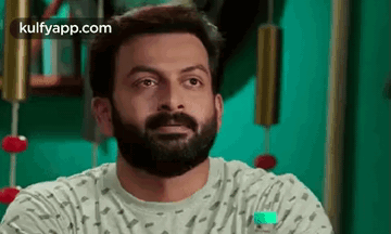 A Man With A Beard Is Sitting In Front Of A Green Wall And Looking At The Camera . GIF