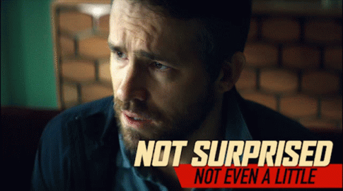 A Man With A Beard Is Sitting In Front Of A Sign That Says Not Surprised GIF