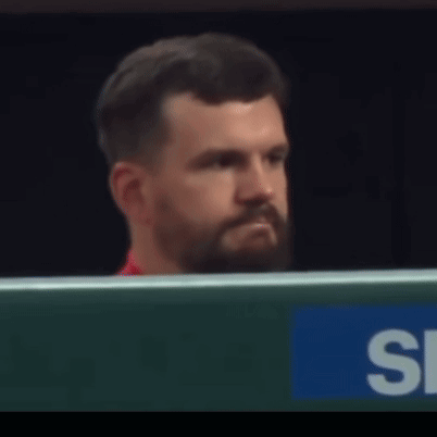 A Man With A Beard Is Sitting In Front Of A Sign That Says S. GIF