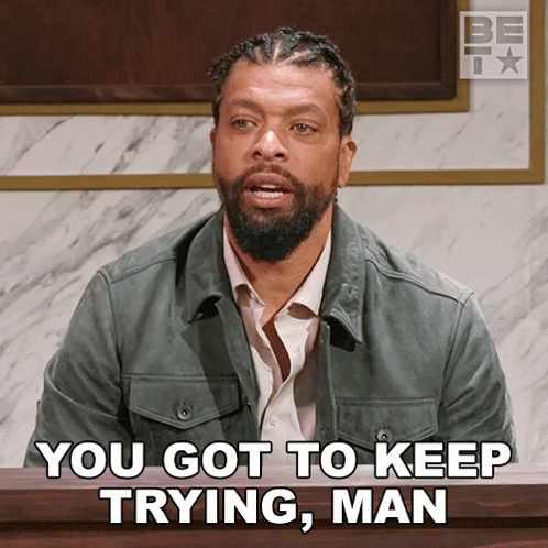 A Man With A Beard Is Sitting In Front Of A Sign That Says " You Got To Keep Trying Man " GIF