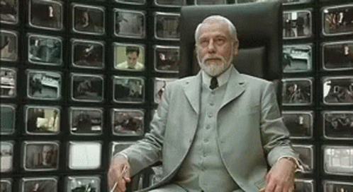 A Man With A Beard Is Sitting In Front Of A Wall Of Televisions GIF