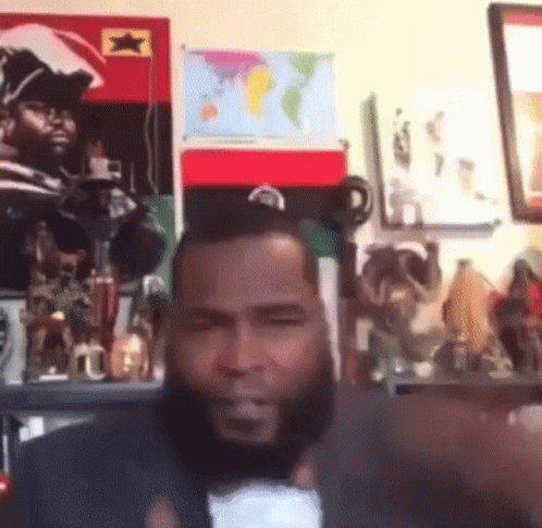 A Man With A Beard Is Sitting In Front Of A Wall With A Map Of The World . GIF