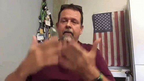 A Man With A Beard Is Sitting In Front Of An American Flag And Making A Hand Gesture . GIF