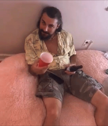 a man with a beard is sitting on a bed holding a beer and a cup