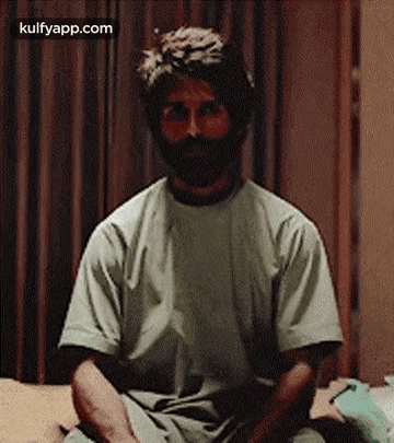 A Man With A Beard Is Sitting On A Bed Wearing A T-Shirt . GIF