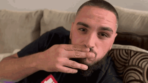 A Man With A Beard Is Sitting On A Couch And Covering His Mouth With His Hand GIF