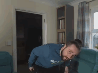 A Man With A Beard Is Sitting On A Couch In A Living Room With A Bookshelf In The Background . GIF
