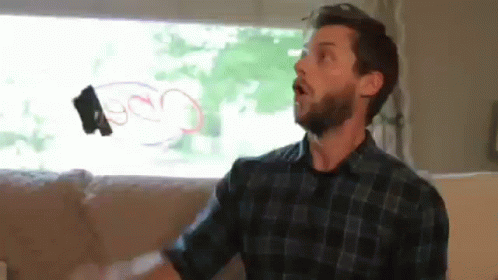 A Man With A Beard Is Sitting On A Couch In Front Of A Window Holding A Remote Control In His Hand . GIF
