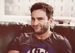 A Man With A Beard Is Sitting On A Couch Wearing A T-Shirt That Says G On It . GIF