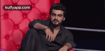 A Man With A Beard Is Sitting On A Red Couch With His Legs Crossed . GIF