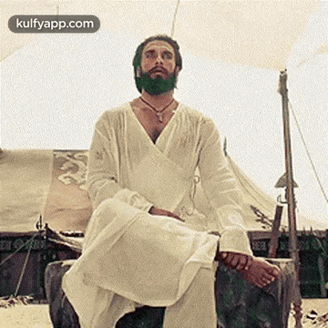 A Man With A Beard Is Sitting On A Rock In Front Of A Tent On The Beach . GIF
