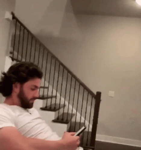 A Man With A Beard Is Sitting On A Staircase Looking At His Phone . GIF
