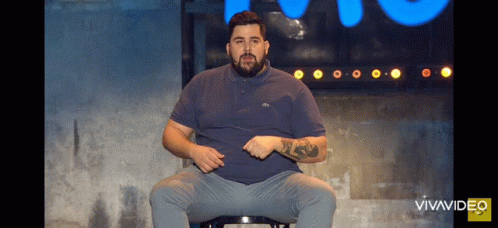 A Man With A Beard Is Sitting On A Stool In Front Of A Blue Sign That Says Vivavideo GIF