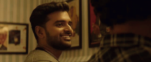A Man With A Beard Is Smiling In Front Of A Wall With Pictures On It GIF