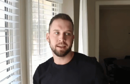 A Man With A Beard Is Smiling In Front Of A Window With Blinds GIF
