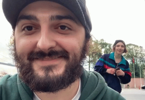 A Man With A Beard Is Smiling While A Woman Stands Behind Him Holding A Cell Phone GIF