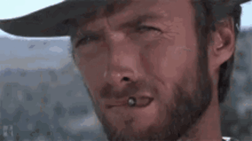 A Man With A Beard Is Smoking A Cigarette And Wearing A Cowboy Hat . GIF