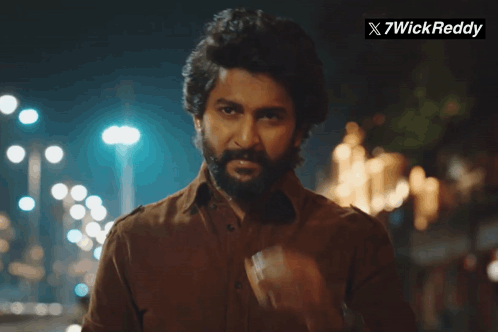 A Man With A Beard Is Smoking A Cigarette In Front Of A Sign That Says X7wick Reddy GIF