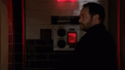 A Man With A Beard Is Standing In A Dark Room With A Red Light Behind Him . GIF
