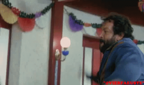 A Man With A Beard Is Standing In A Room With A Lamp And Decorations On The Wall . GIF