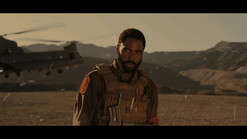 A Man With A Beard Is Standing In Front Of A Helicopter And Mountains GIF